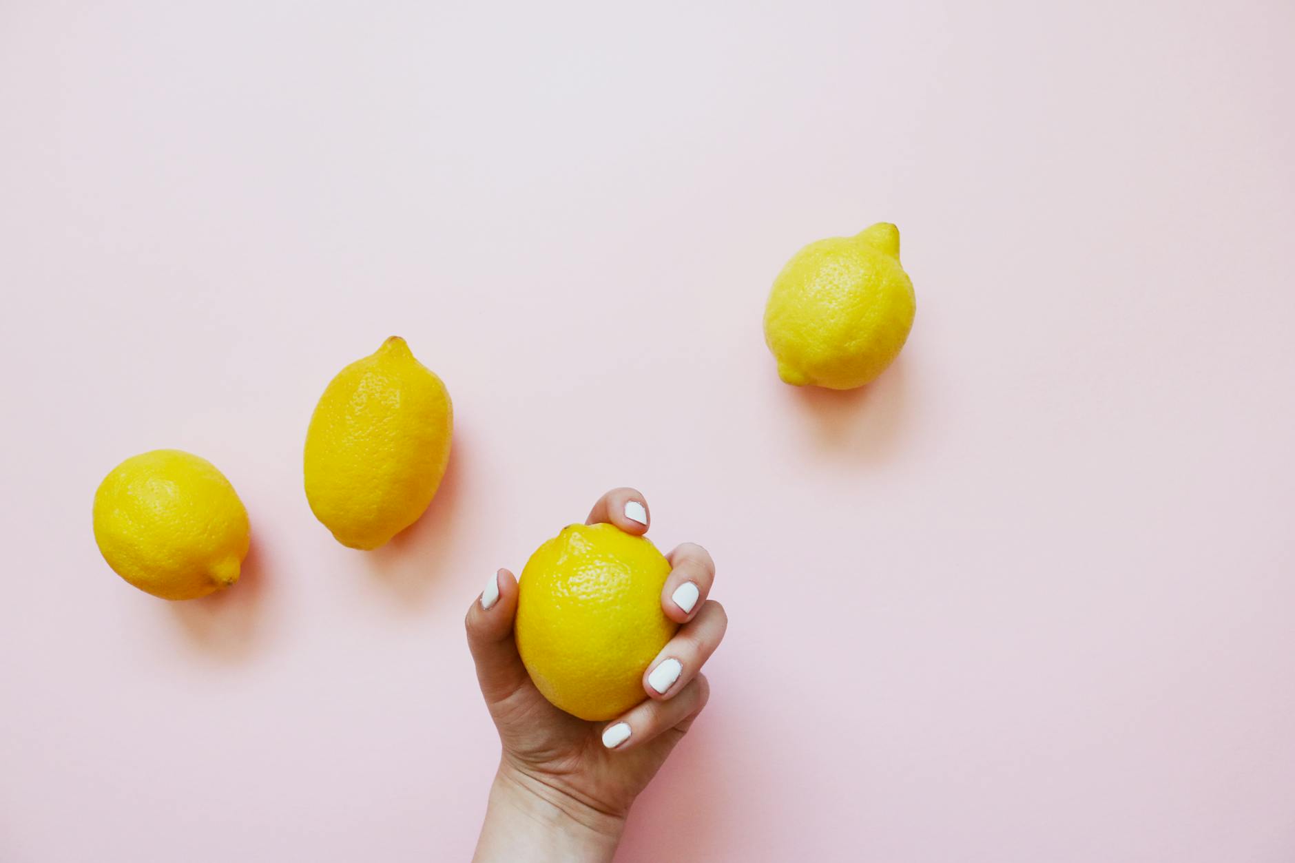 A hand with white nails holding a lemon on a soft pink background, symbolizing gentle relief and natural tension release