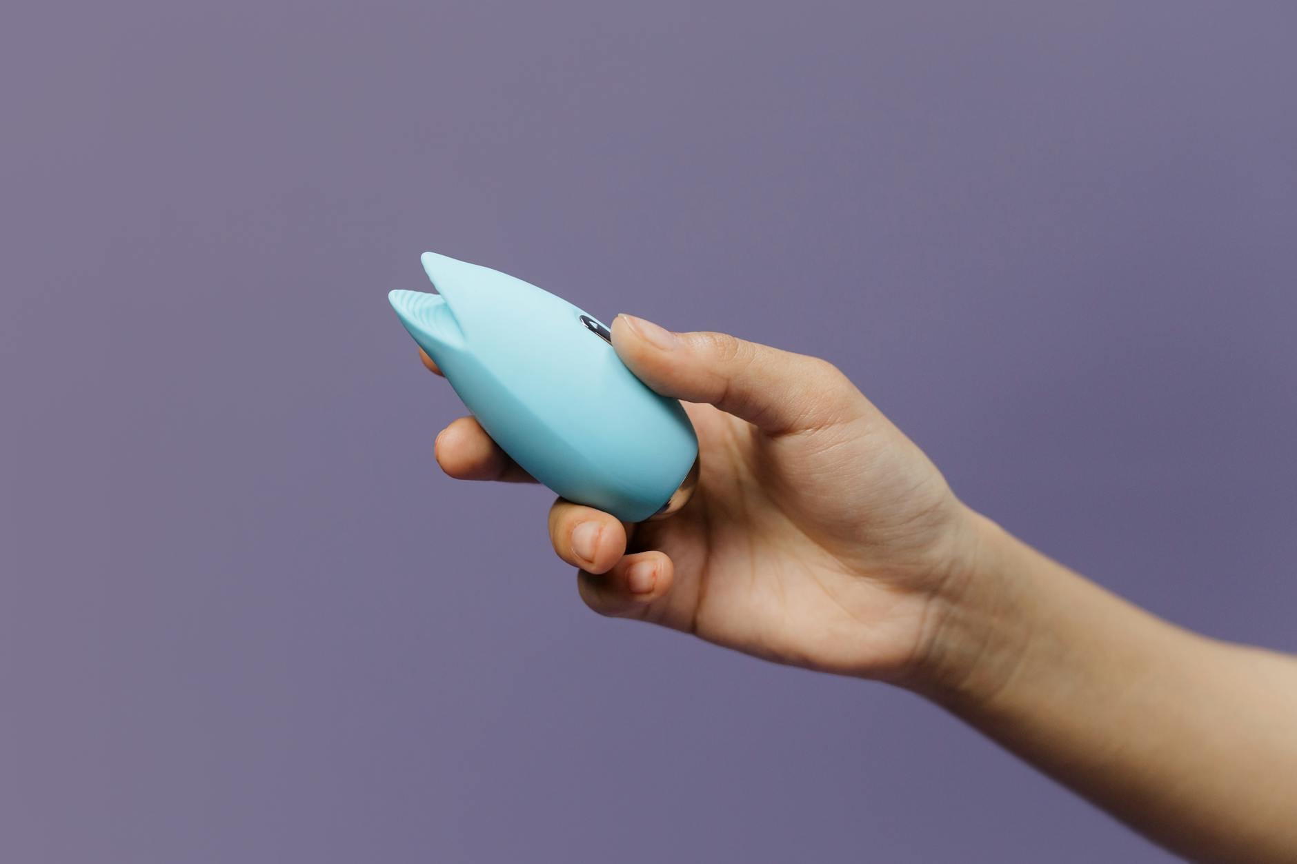 Hand holding a blue silicone clitoral vibrator against a purple background, representing sexual wellness and pleasure